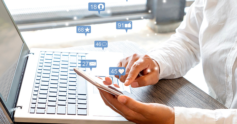 Business-owner-at-a-laptop-looking-at-a-smart-phone-that-has-Facebook-comment-icons-overlaid-above-it.