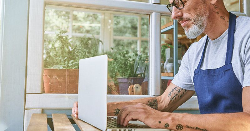Homme portant des lunettes et un tablier, assis à un bureau dans sa petite entreprise et utilisant son ordinateur portable.