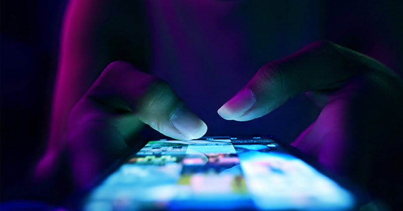 A woman's-thumbs-poised-over-a-light-up-smart-phone-in-a-dark-room.