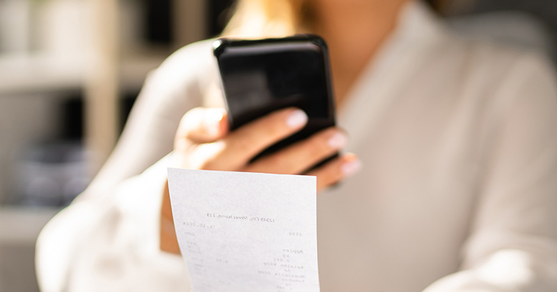 Young-business-woman-holding-up-a-paper-receipt-to-scan-with-her-smart-phone.
